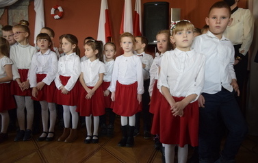 Przedszkolaki ubrane w białe koszule i czerwone sp&oacute;dniczki, śpiewają piosenkę