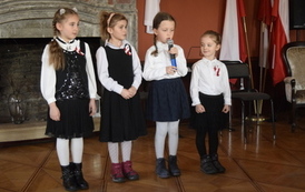 Cztery małe dziewczynki w galowych strojach, jedna z mikrofonem recytuje wiersz