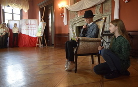 Dwie dziewczyny przebrane za mężczyzn odgrywają scenkę. Jedna siedzi na fotelu i czyta książkę a druga z nią rozmawia. W tle dekoracja patriotyczna.