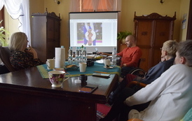 Trzy kobiety i mężczyzna siedzą przy stole. Mężczyzna z laptopa wyświetla na dużym ekranie prezentację multimedialną.