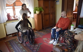 Troje ludzi. Kobieta i mężczyzna siedzą na przeciwko siebie na w&oacute;zkach inwalidzkich. Druga kobieta stoi pochylona za w&oacute;zkiem inwalidzkim na kt&oacute;rym siedzi pierwsza kobieta.