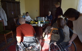 Grupa ludzi siedzi przy stole, rozmawiają. Dwoje z nich, kobieta i mężczyzna, siedzą na w&oacute;zkach inwalidzkich.