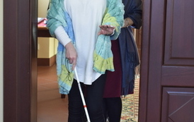 Do pomieszczenia wchodzi kobieta w goglach na oczach a w ręce ma białą laskę. za nią wchodzą dwie kobiety.