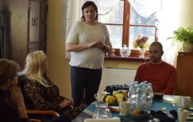 w pokoju kobieta i mężczyzna siedzą przy stole i słuchają kobiety kt&oacute;ra przemawia na stojąco. 